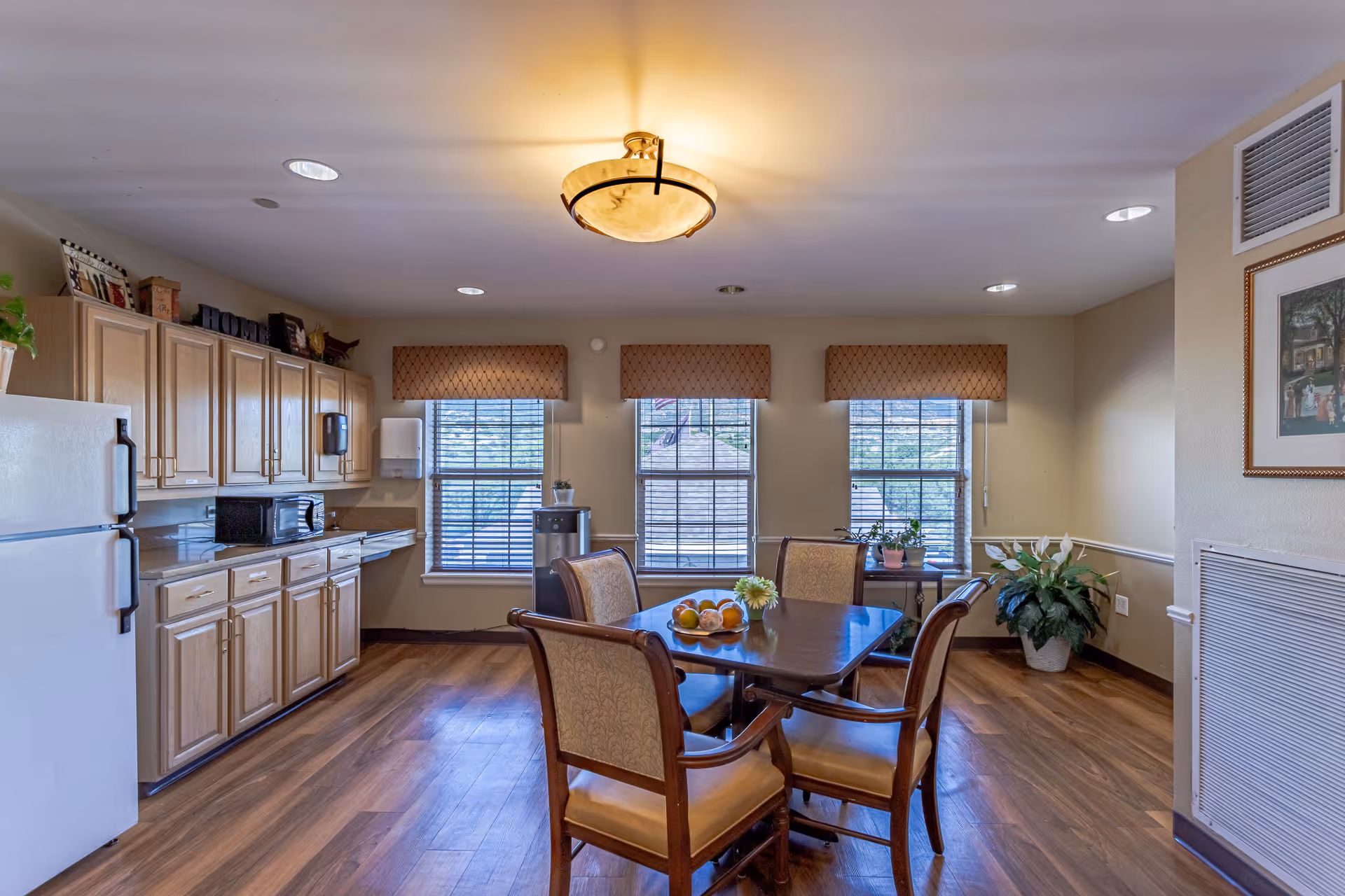 A bright kitchen and dining area with wooden floors, beige walls, and three windows with patterned valances. The kitchen has light wood cabinets, a white refrigerator, a microwave, and a water cooler. A wooden dining table with four upholstered chairs is centered in the room, decorated with a small flower arrangement and a bowl of fruit. There are also some potted plants and framed artwork on the walls.