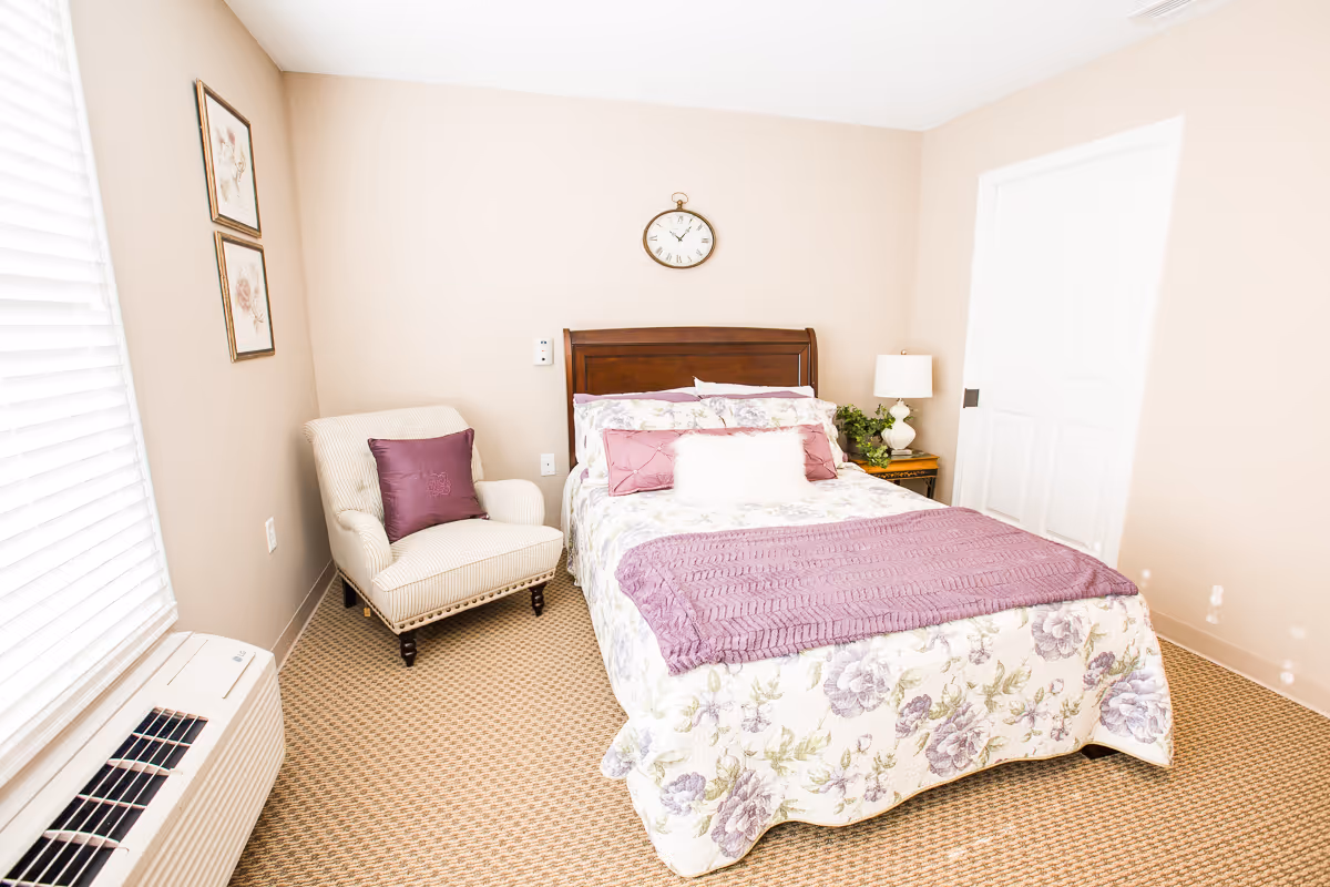 A cozy bedroom with a floral-patterned bedspread and purple accents, including pillows and a throw blanket. There is a cushioned armchair with a purple pillow next to a window with blinds. A wooden headboard is against the wall with a round clock above it. A nightstand with a lamp and a small plant is beside the bed. The room has beige walls and carpeted flooring.