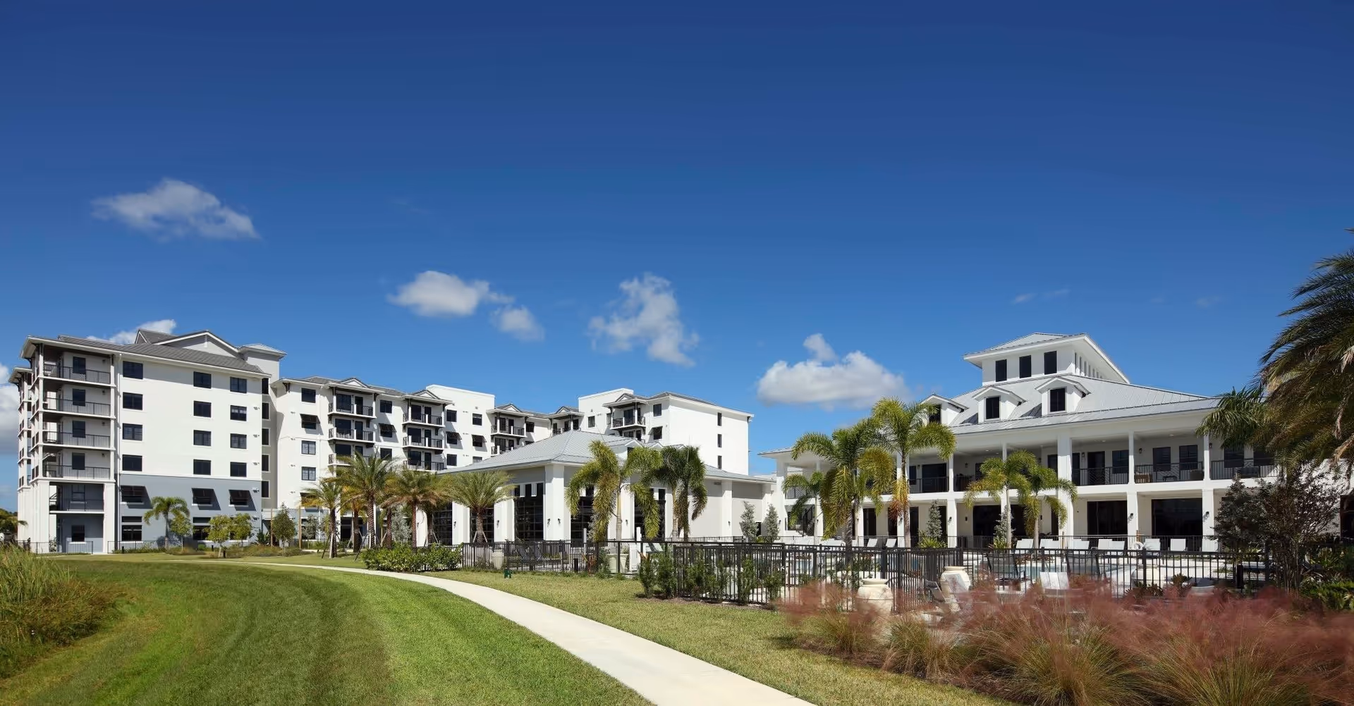 Exterior view of Wellington Bay senior living facility showing multiple white buildings with balconies, palm trees, a well-maintained lawn, and a clear blue sky with a few clouds.