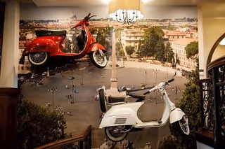 Two vintage scooters, one red and one white, mounted on a wall inside a building with a large mural of a European city square in the background. The setting appears to be an interior space with a chandelier and decorative railing visible.
