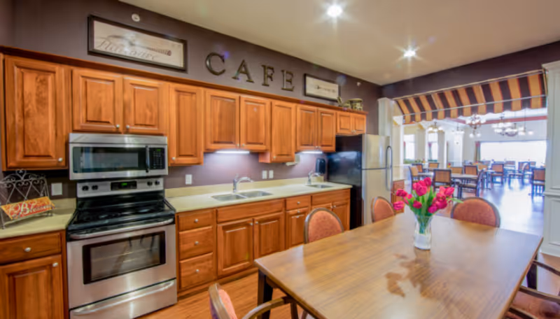 Open communal kitchen with wooden cabinets, stainless stove and sink, and a dining table with a vase of pink flowers overlooking a dining area.