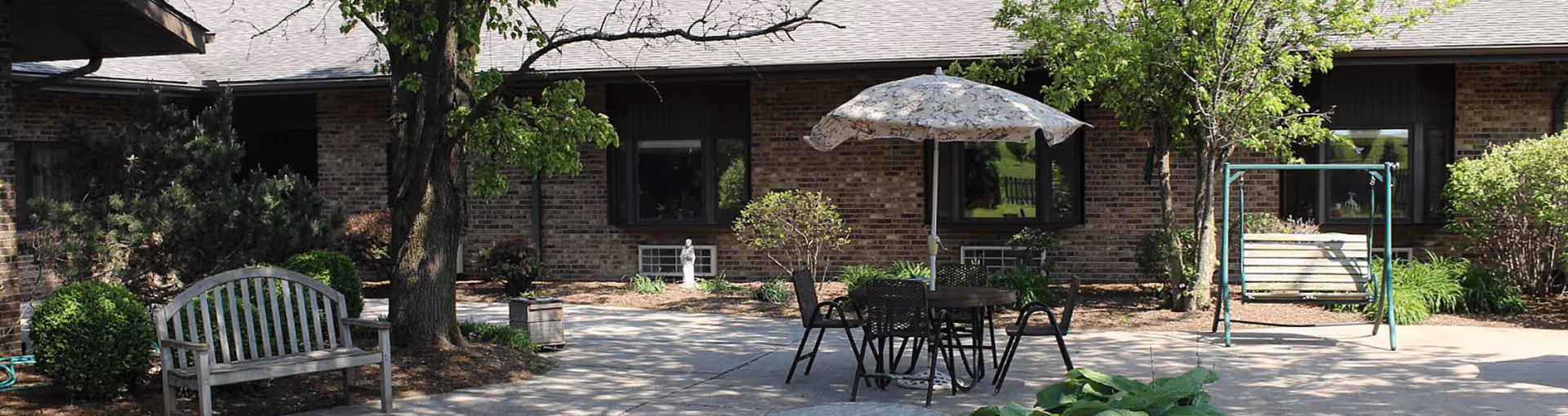 Outdoor patio area at Mason City Area Nursing Home with a wooden bench, a round table with four chairs and a floral umbrella, a metal swing, trees, shrubs, and a brick building in the background.