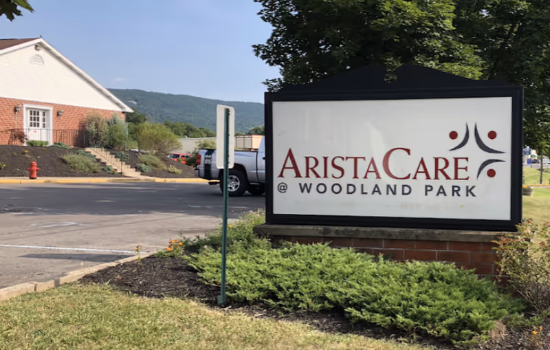 A landscaped lawn and parking area with a sign reading 'AristaCare @ Woodland Park' in front of the facility building and nearby hills.