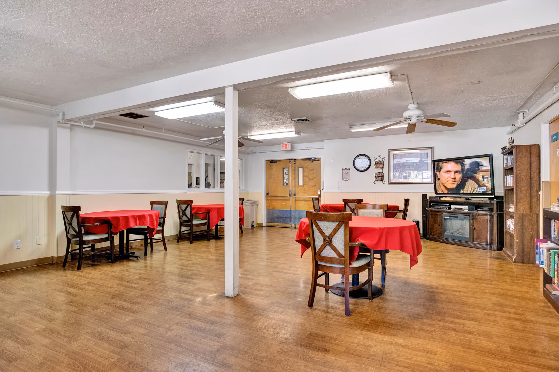 Large common dining/activity room with round tables covered in red tablecloths, chairs, a television and bookshelves.