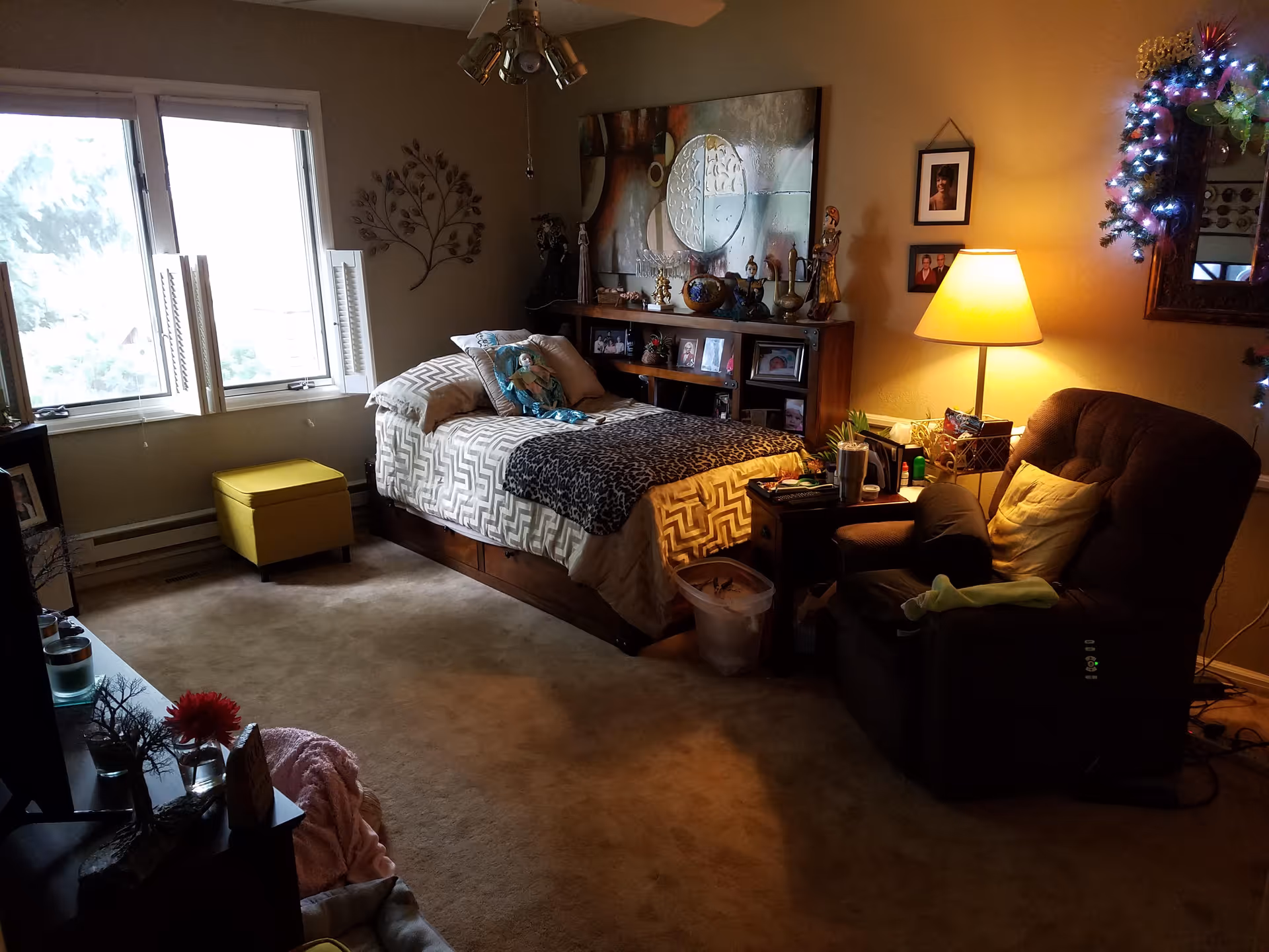 A cozy bedroom with a single bed featuring patterned bedding and a stuffed doll. The room has a large window with white shutters, a yellow ottoman, a brown recliner chair with a pillow, and a side table with a lamp. The walls are decorated with framed photos, a large abstract painting, and a metal tree wall art. There is a mirror with a wreath of lights on the right side of the room.