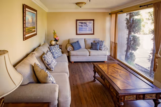 A cozy living room with two beige sofas adorned with patterned and solid blue cushions, a wooden coffee table with intricate designs, a table lamp on a side table, framed artwork on the walls, and a large window with beige curtains letting in natural light and showing greenery outside.