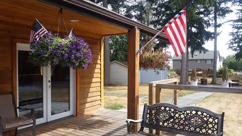 A wooden porch with hanging flower baskets and an American flag. There is a decorative metal bench on the porch, and a grassy yard with trees and houses in the background.
