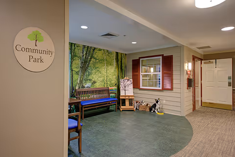 Indoor seating area labeled Community Park with a wooden bench with blue cushions, a small table, and decorative items including a basket with stuffed animals and a framed picture. The wall behind the bench features a large mural of a green forest scene. The area has a mix of green flooring and carpet, with beige walls and ceiling lights.
