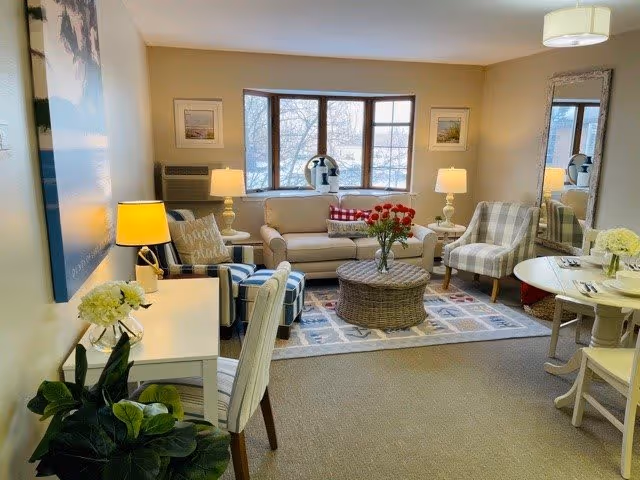A cozy living room with a beige sofa, two patterned armchairs, a round wicker coffee table with a vase of red flowers, two table lamps on side tables, a large window with a view of trees, a white desk with a chair and a plant, and a round dining table with two chairs. The room is warmly lit and decorated with a large wall art and a mirror.