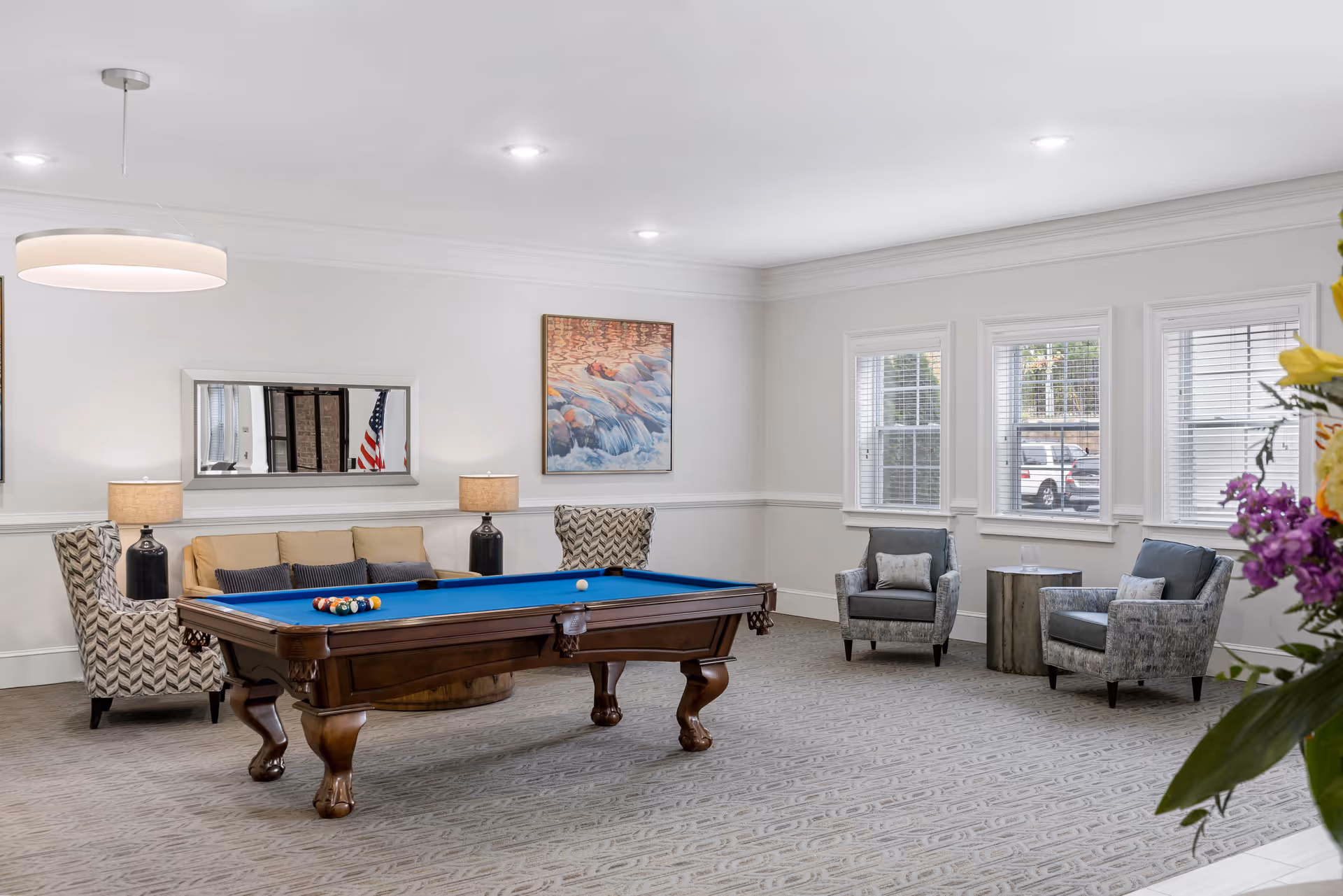 A bright and spacious room with a blue felt pool table in the center, surrounded by comfortable seating including a beige sofa with cushions, two patterned armchairs, and two gray armchairs near three windows with white blinds. The room has white walls, a large mirror, a painting, two table lamps, and a modern ceiling light fixture. A vase with colorful flowers is partially visible on the right side.