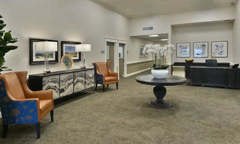 A spacious and well-lit interior common area with a round wooden table in the center holding a white pot with white orchids. To the left, there are two brown leather armchairs with blue patterned backs, and a sideboard with two table lamps, framed artwork, and a decorative plate. On the right side, there is a dark wooden reception desk with three framed botanical prints on the wall behind it. The carpet is beige, and the walls are painted in neutral tones.