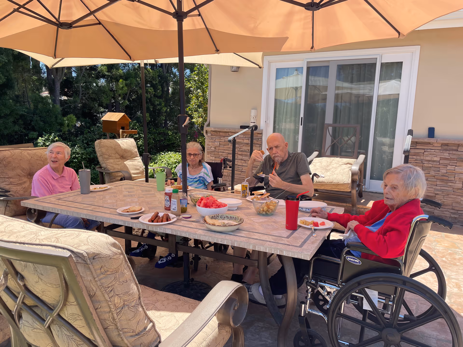Four elderly individuals sitting around a large outdoor table under a beige patio umbrella. The table has various plates of food including watermelon, sausages, and snacks. Two of the individuals are in wheelchairs. The setting is a patio area with cushioned chairs and greenery in the background.