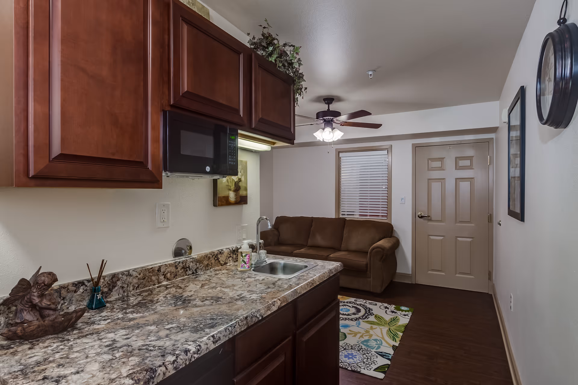 Small assisted-living suite kitchenette with a marble countertop, sink and cabinets opening onto a living area with a brown sofa and ceiling fan.