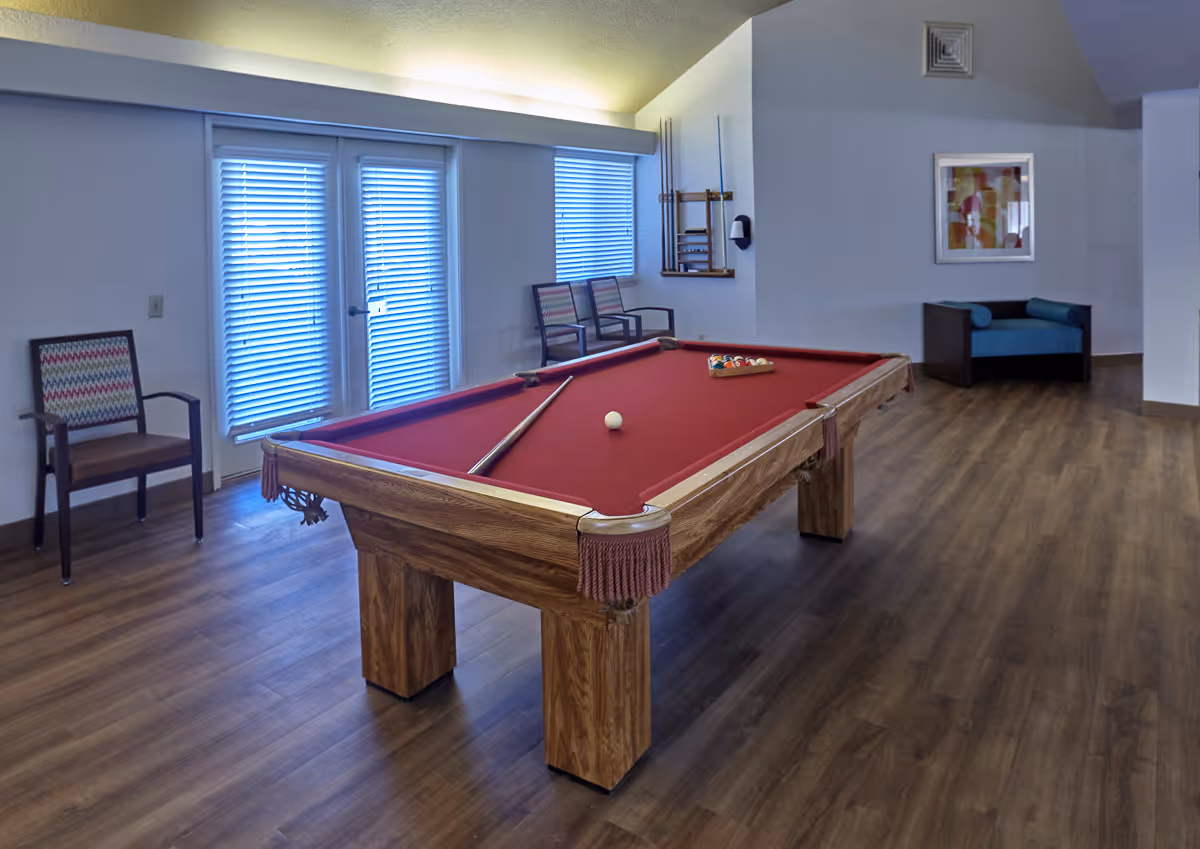 A recreational room with a red felt pool table in the center, a cue stick resting on the table, and a rack of billiard balls. The room has wooden flooring, two chairs with patterned cushions near a set of glass doors with blinds, a wall-mounted pool cue rack, and a blue cushioned armchair against the wall with a framed abstract artwork above it.