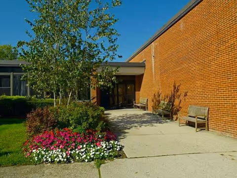 Walkway leading to a brick building entrance with benches, a flowerbed, and trees under a clear blue sky.