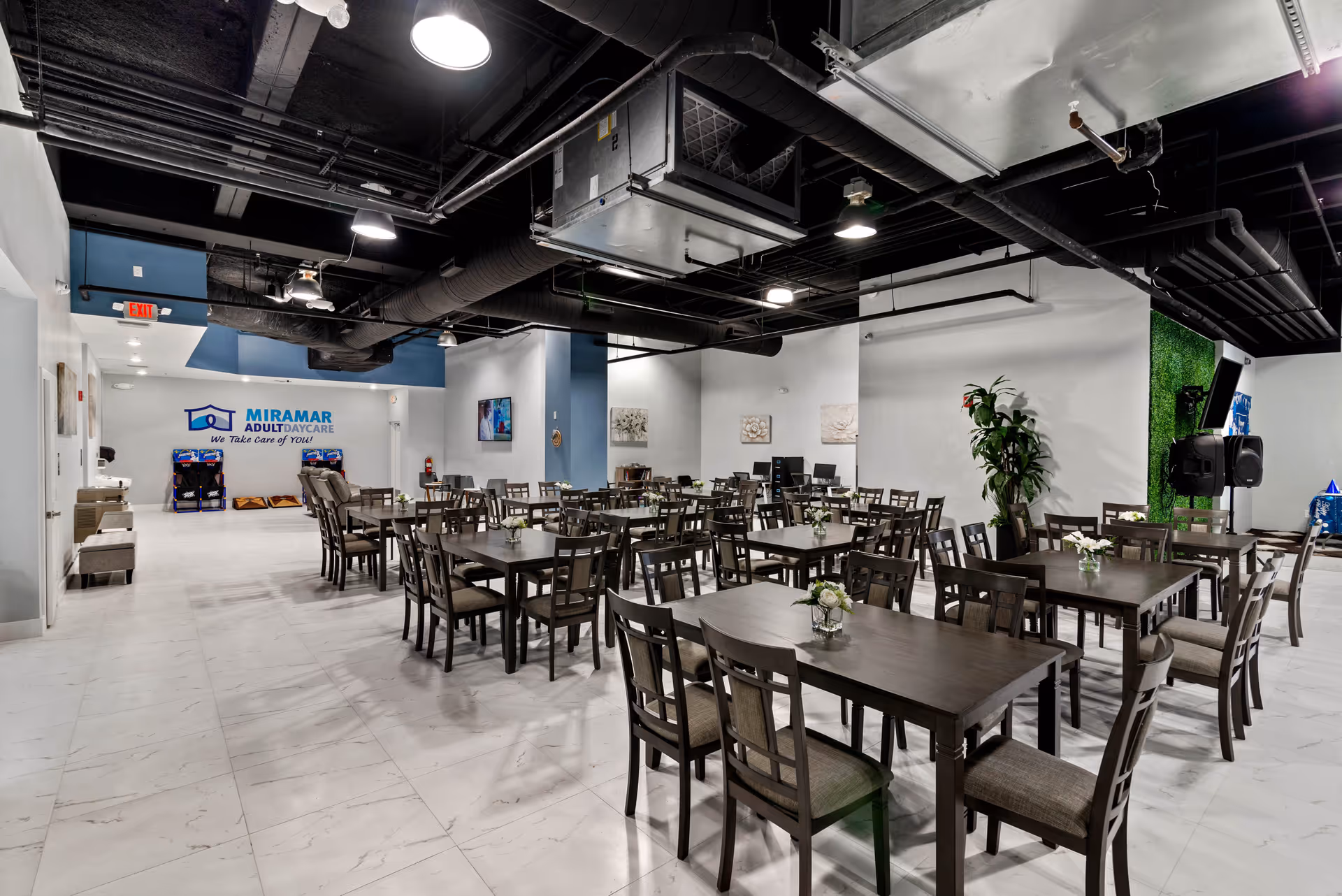 A spacious dining area in Miramar Adult Day Care with multiple dark wooden tables and chairs arranged neatly. Each table has a small flower arrangement. The room has white tiled floors, white walls with some blue accents, and an exposed black ceiling with visible ducts and lights. There is a sign on the far wall that reads 'Miramar Adult Day Care We Take Care of YOU!' and some arcade machines near the back wall.