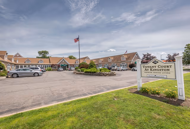 Exterior view of Tiffany Court at Kingston, a single-story senior living facility with a parking lot in front, an American flag on a flagpole, and a landscaped area with bushes and trees under a partly cloudy sky.
