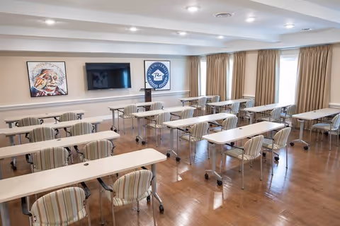 A well-lit classroom or meeting room with multiple white tables and striped cushioned chairs on wheels arranged in rows facing a wall-mounted flat screen TV. The room has wooden flooring, beige curtains covering the windows, and two framed artworks on the wall, one featuring a portrait and the other a circular emblem.
