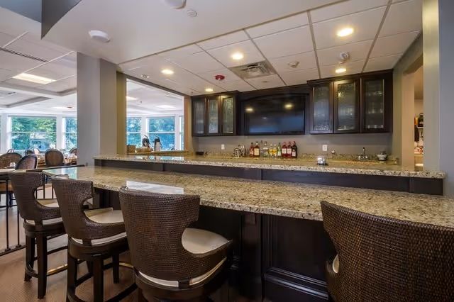 Interior view of a bar area in a senior living facility with a granite countertop, four wicker bar stools with cushions, a wall-mounted flat screen TV, and shelves with glass doors holding bottles of liquor. In the background, there is a dining area with tables and chairs near large windows letting in natural light.