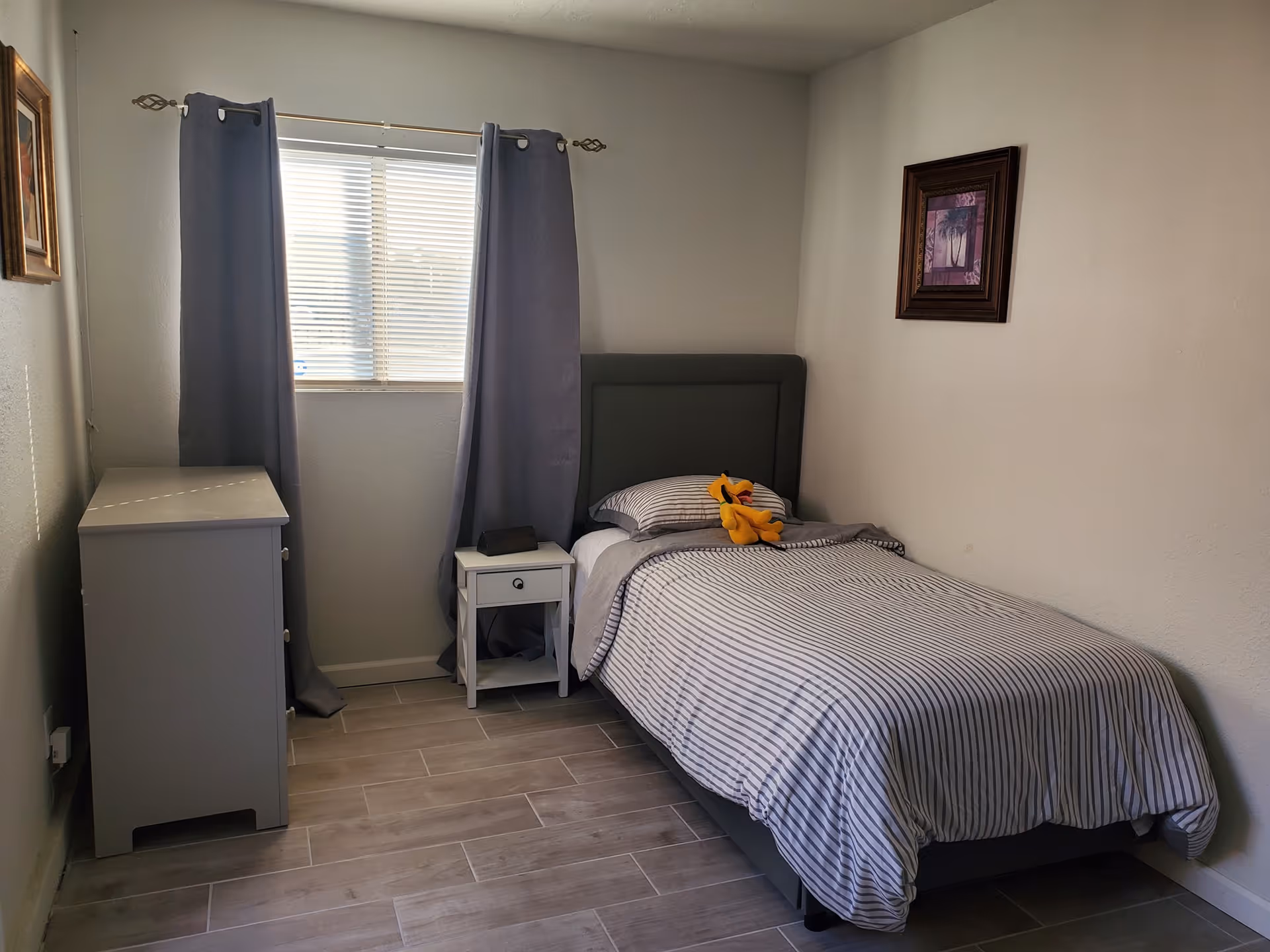 A small bedroom with a single bed covered in striped bedding and a plush toy on the pillow. There is a window with gray curtains, a white nightstand with a small item on it, a gray dresser, and a framed picture on the wall.
