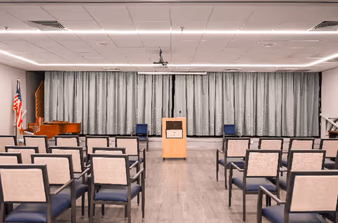 An empty meeting or presentation room with rows of chairs facing a wooden podium in the center. Behind the podium is a closed curtain covering the entire back wall. To the left, there is an American flag and a piano. The room has a modern ceiling with recessed lighting and a projector mounted in the center.