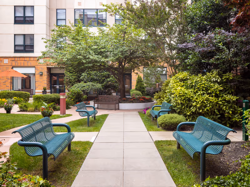 Outdoor courtyard area with green metal benches along a paved walkway, surrounded by well-maintained bushes, trees, and flowering plants, with a multi-story building in the background.