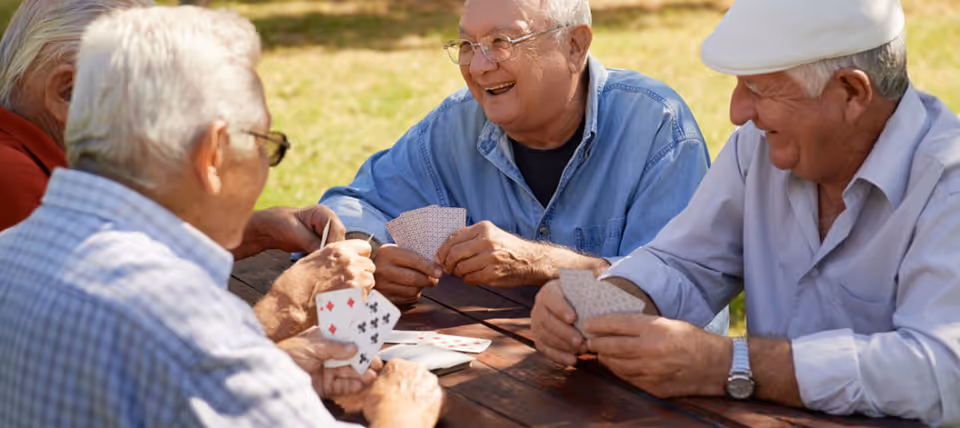 A group of elderly men sitting outdoors at a wooden picnic table playing cards and smiling, enjoying a sunny day.