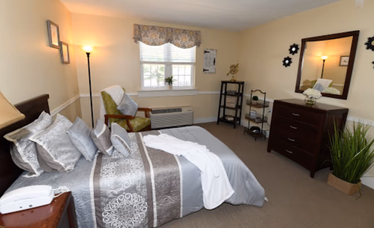 A cozy bedroom in a senior living facility featuring a neatly made bed with multiple decorative pillows and a patterned bedspread. There is a green armchair with a pillow and a white throw draped over it near a window with blinds and a valance. A floor lamp provides warm lighting. On the opposite wall, there is a dark wooden dresser with a mirror above it, decorated with a vase of white flowers. Next to the dresser is a small shelving unit with decorative items and a tall green plant in a basket on the floor.
