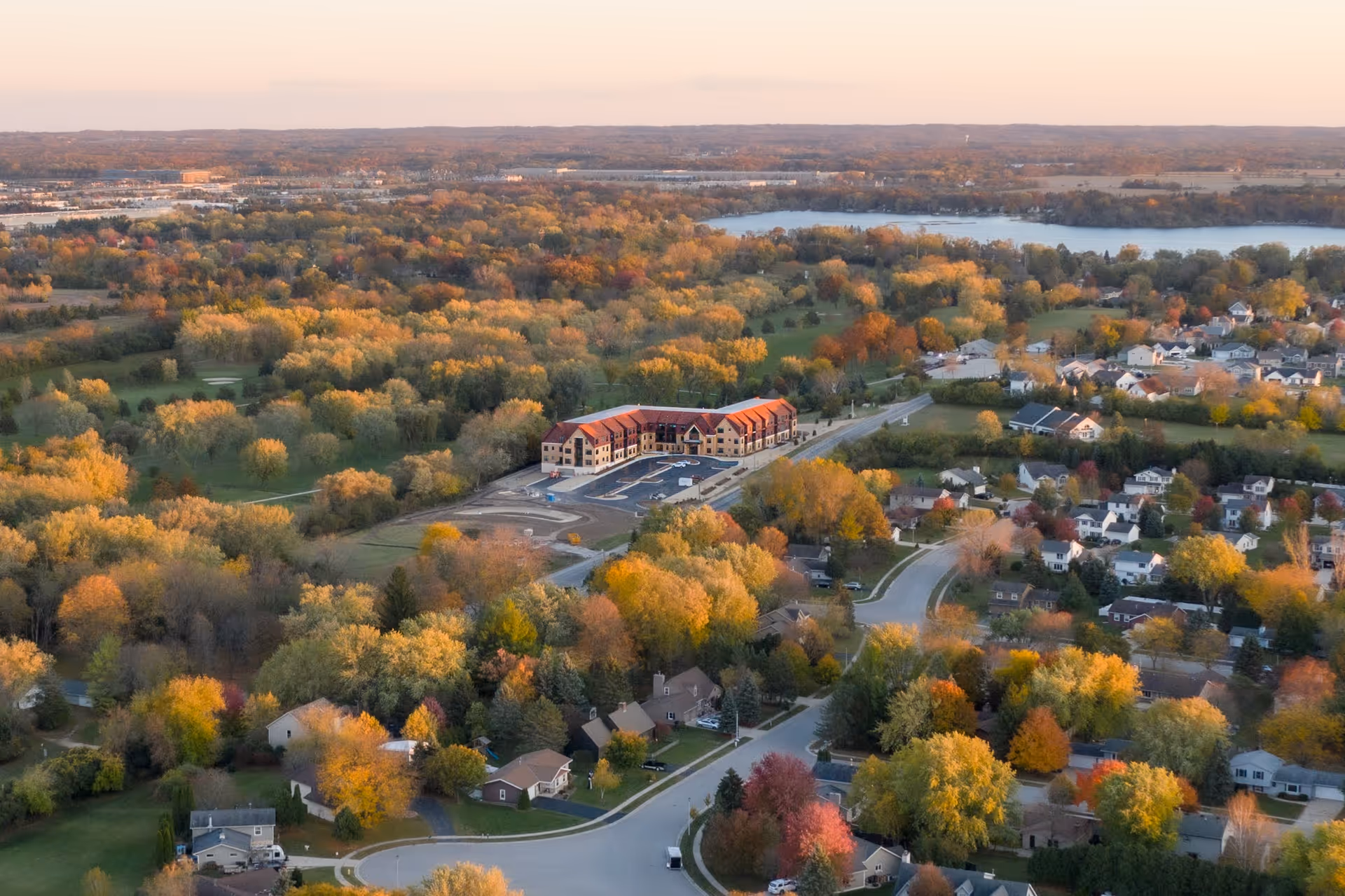 Aerial view of a residential area with a large building complex surrounded by trees with autumn foliage. The landscape includes houses, roads, and a body of water in the distance under a clear sky.