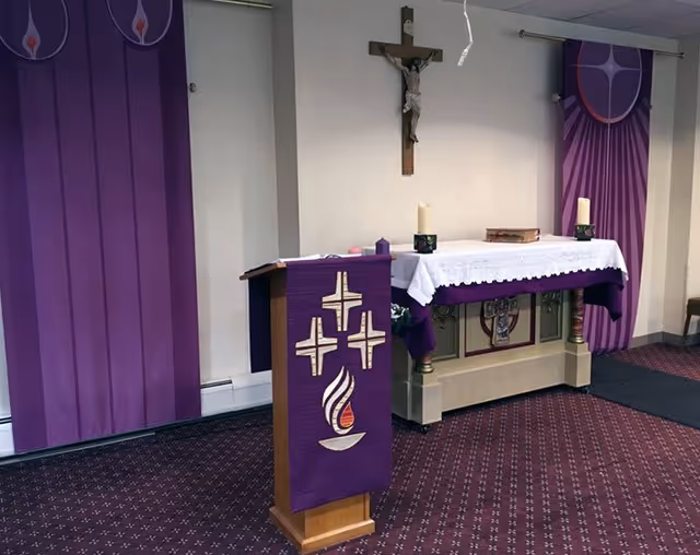 Interior view of a chapel or prayer room with a wooden podium draped in a purple cloth featuring crosses and a flame symbol. Behind the podium is an altar covered with a white cloth and purple accents, holding two candles and a book. A crucifix is mounted on the wall above the altar, and purple banners with religious symbols hang on the walls. The floor is carpeted in a purple pattern.