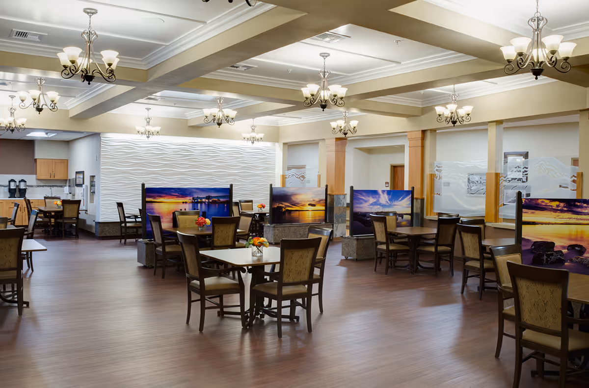 Spacious dining room with multiple tables and chairs, scenic decorative partition panels, chandeliers, and a serving area in the back.