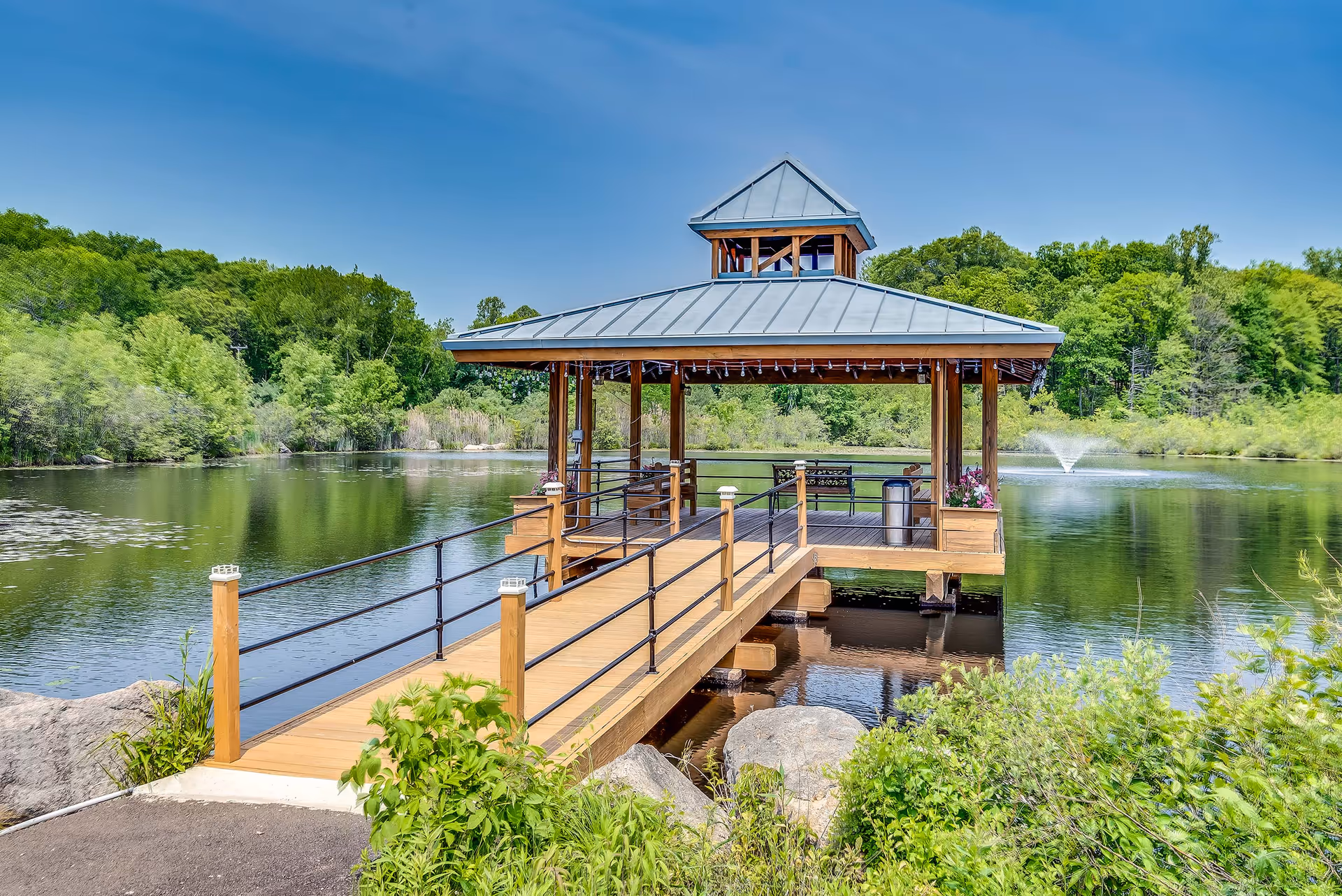 A wooden gazebo with a metal roof extends over a calm pond surrounded by lush green trees. The gazebo is accessible by a wooden walkway with railings and has benches and flower pots inside. A water fountain sprays water in the pond in the background under a clear blue sky.
