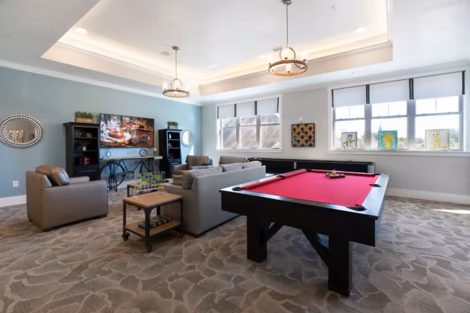 A bright and spacious common room with a red pool table in the foreground, a gray sofa set with armchairs, a TV mounted on a light blue wall, decorative mirrors, and large windows letting in natural light. The room has patterned carpet flooring and modern ceiling lights.