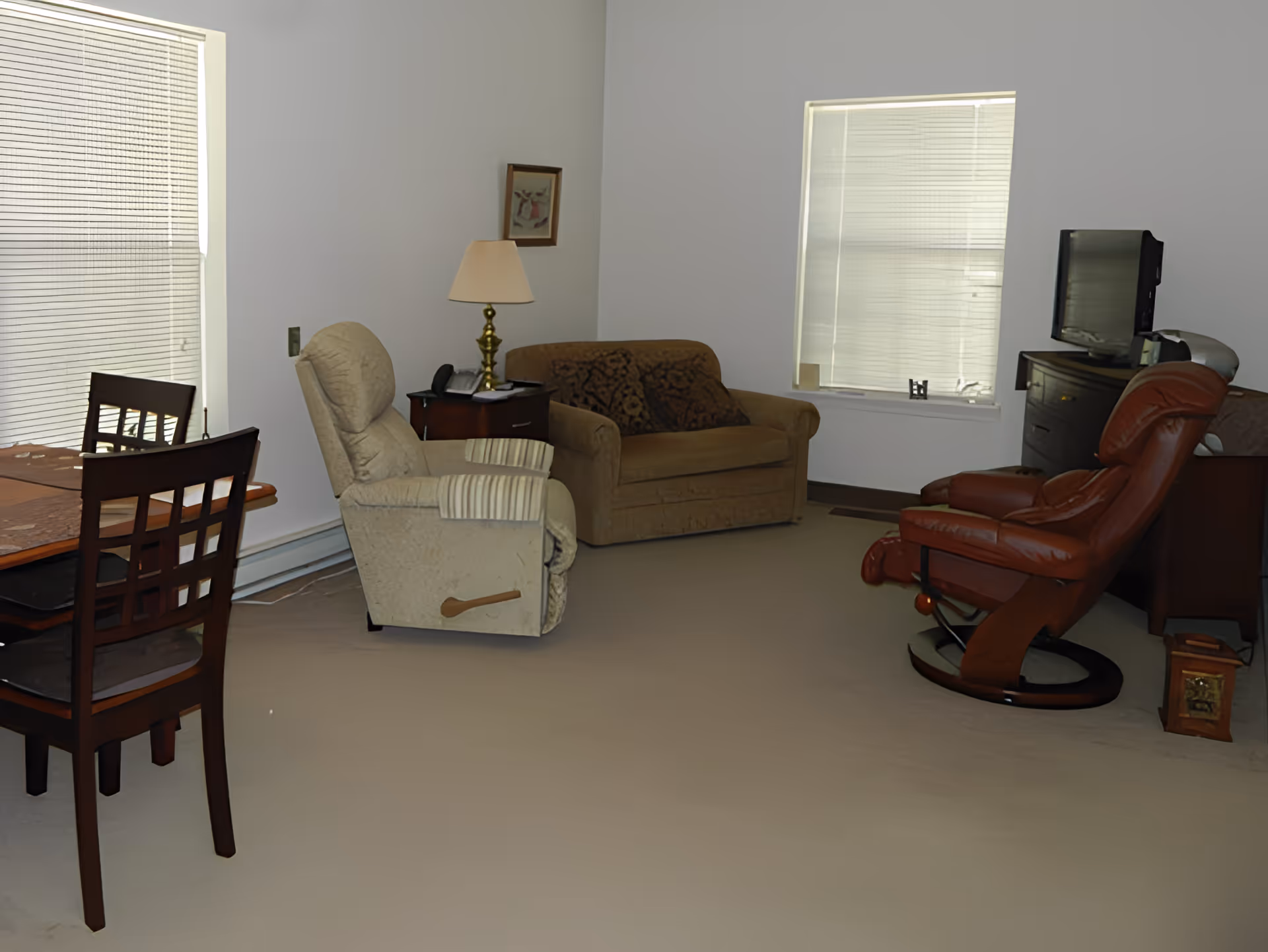 A cozy living room in Aspen Ridge Retirement Village featuring a beige recliner, a brown loveseat with a patterned throw, a wooden side table with a brass lamp and telephone, a wooden dining table with chairs, a red leather swivel chair, a TV on a dark wooden dresser, and two windows with blinds.