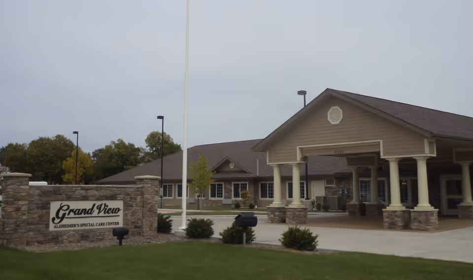 Exterior view of the Grand View Alzheimer's Special Care Center building with a stone sign in front displaying the facility name. The building has a covered entrance supported by columns and is surrounded by a driveway and some greenery.