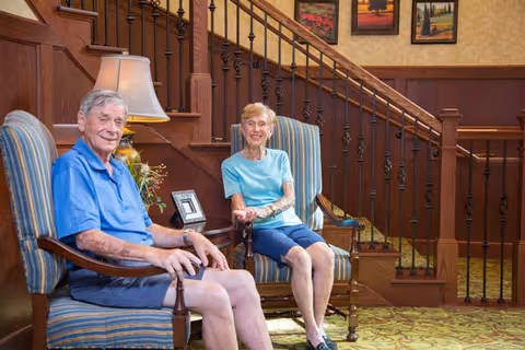 An elderly man and woman sitting on striped armchairs in a warmly decorated common area with wooden staircase and framed artwork on the wall behind them.