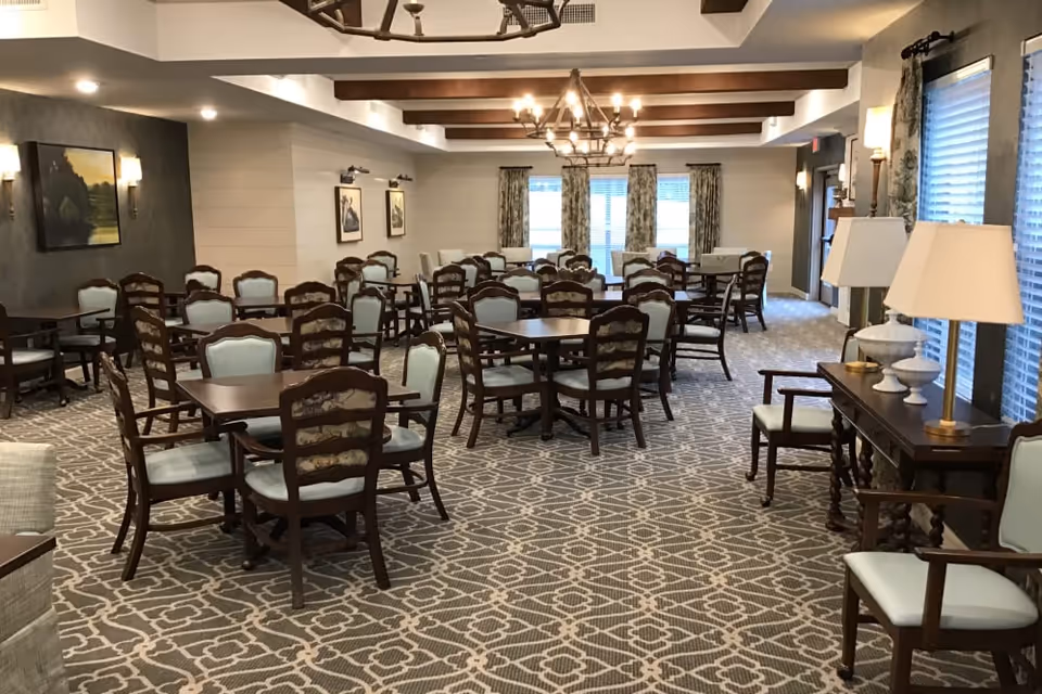 Large, well-lit dining room with multiple wooden tables and upholstered chairs arranged on patterned carpet under chandeliers.