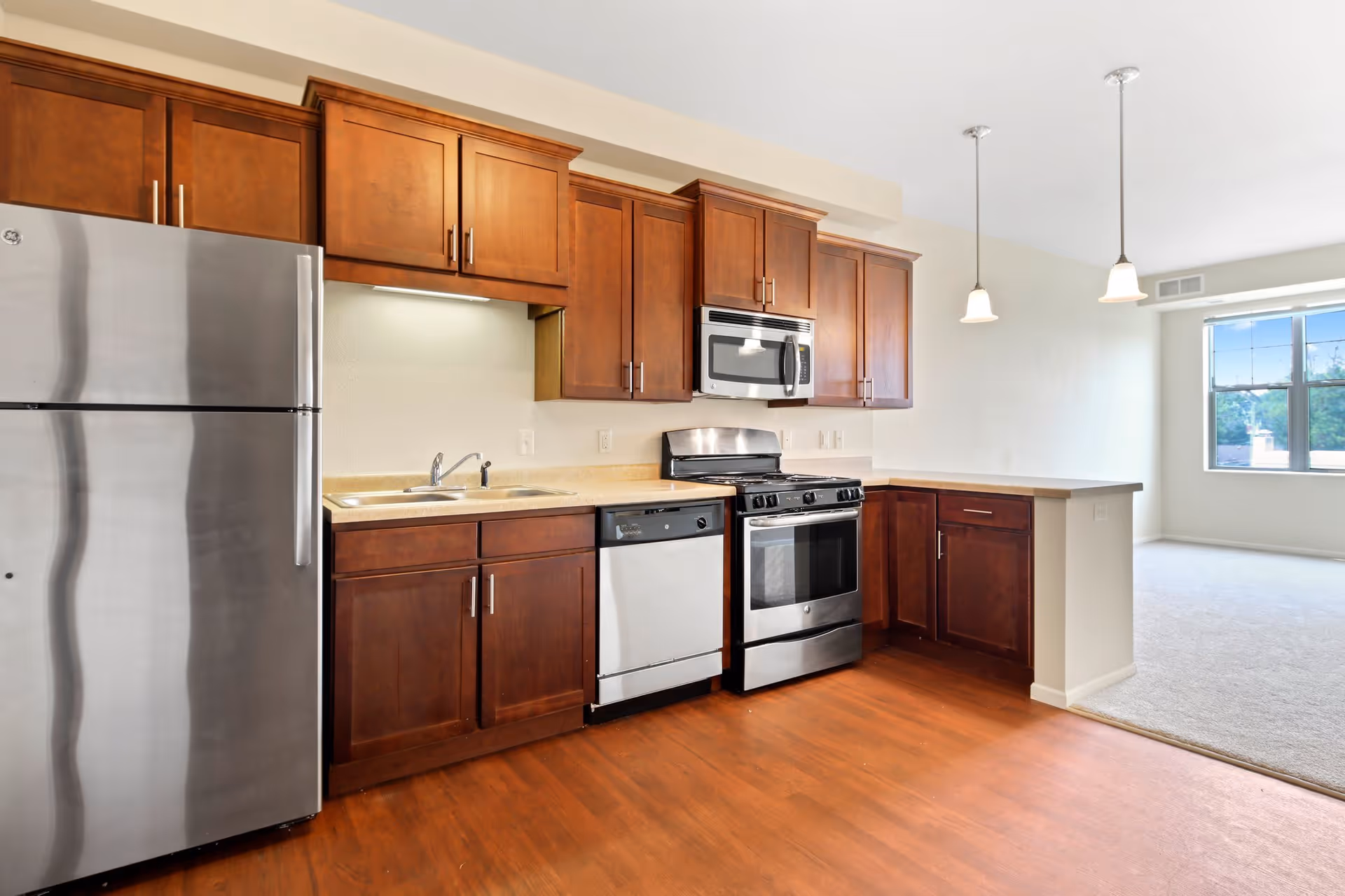 Modern kitchen with stainless steel refrigerator, dishwasher, oven, and microwave. The kitchen has wooden cabinets and a light-colored countertop. The floor is wooden, and there are two pendant lights hanging from the ceiling. An adjacent room with carpeted flooring and a large window is visible in the background.