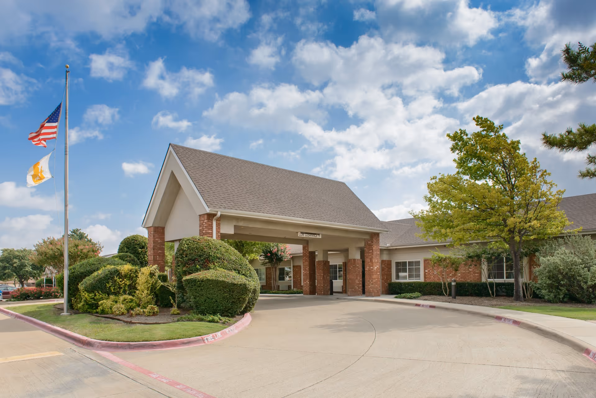 Exterior view of Life Care Center of Plano showing the entrance with a covered driveway, well-maintained bushes and trees, and two flagpoles with the American flag and another flag flying against a partly cloudy sky.