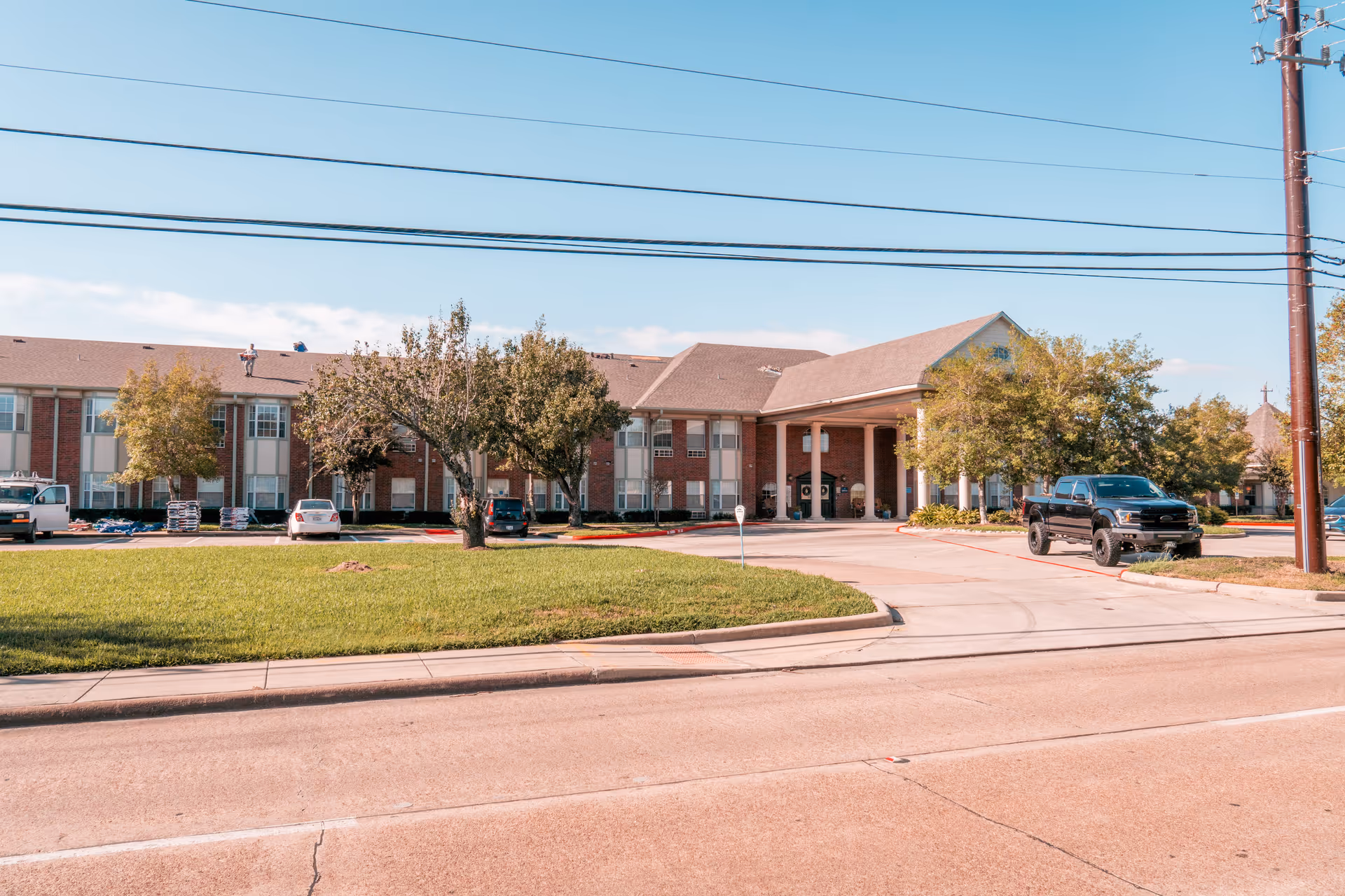 Exterior view of a two-story senior living facility building with a covered entrance supported by columns. There are several trees and parked vehicles in front of the building, with a well-maintained lawn and a clear blue sky overhead.