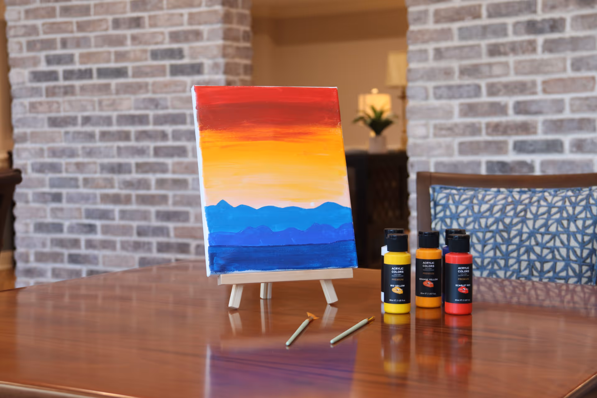 A small colorful canvas on an easel with paint bottles and brushes arranged on a wooden table in a brick-walled interior.