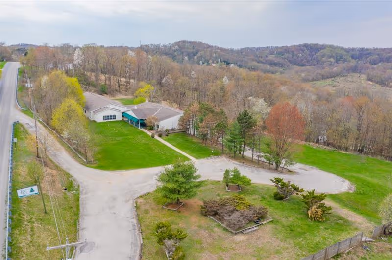 Aerial view of a single-story building surrounded by green lawns and trees with a winding road leading to the entrance, set in a wooded, hilly landscape.