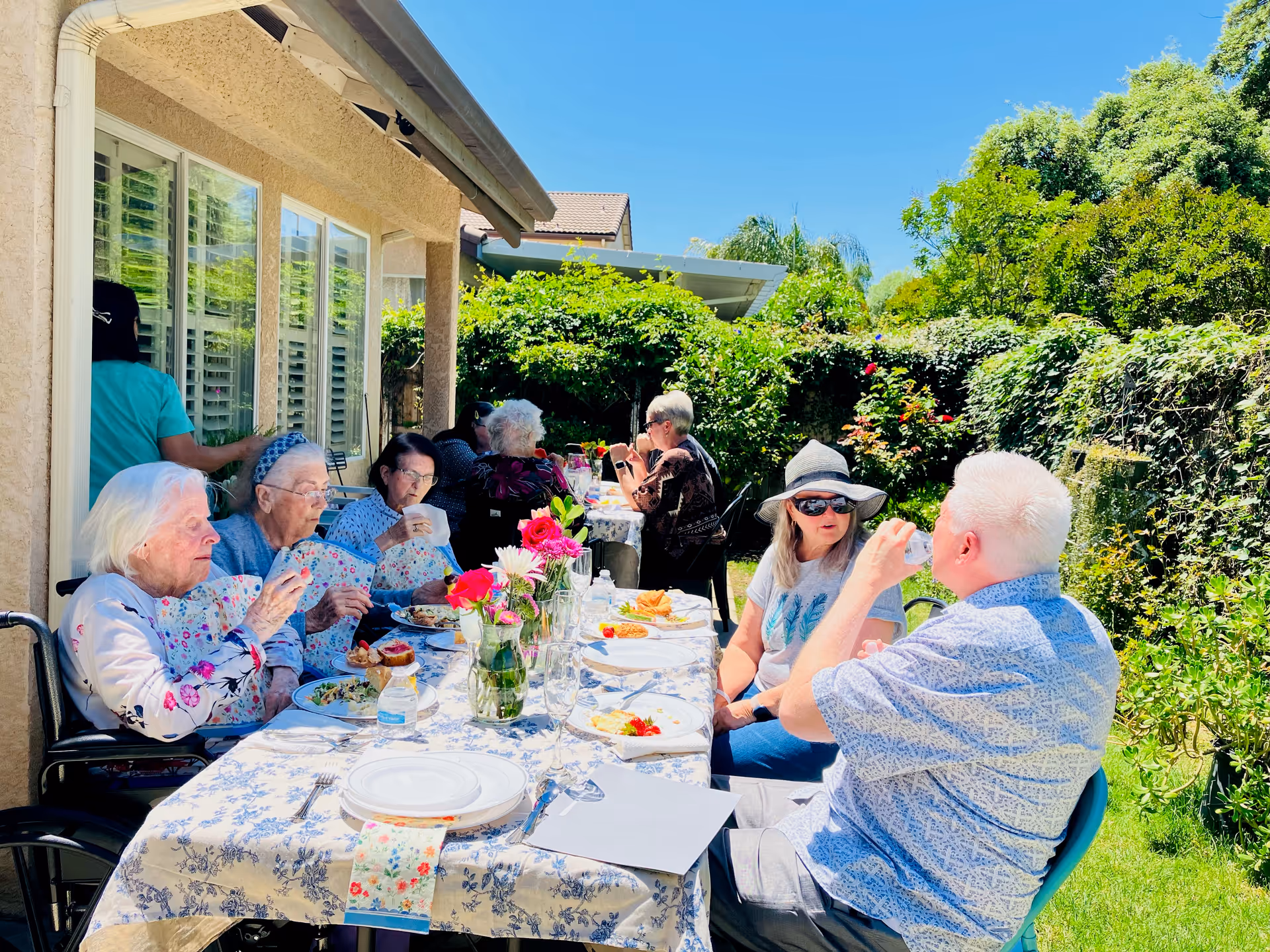A group of elderly people sitting around a long outdoor table covered with a floral tablecloth, enjoying a meal together in a garden area with lush greenery and bright sunlight. Some are eating, drinking, and conversing, with flowers in vases decorating the table.