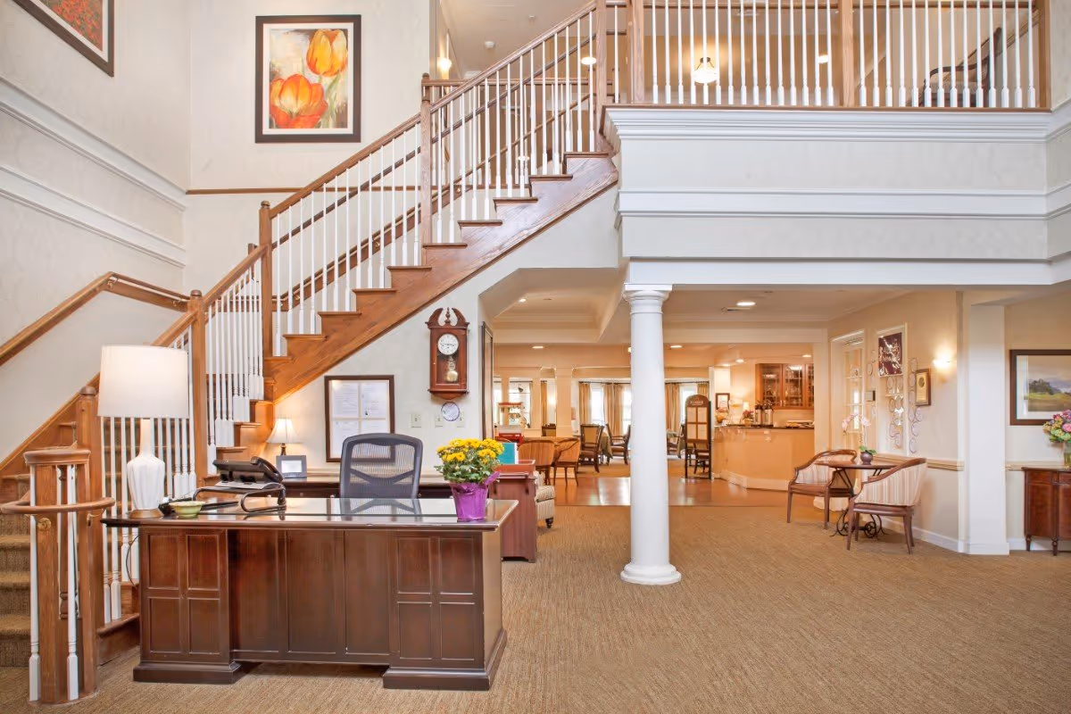 Interior view of a senior living facility lobby with a wooden reception desk, a chair, a lamp, and a potted plant. Behind the desk is a staircase with wooden handrails and white balusters leading to an upper floor. The area is decorated with framed artwork, a wall clock, and has beige walls and carpeted flooring. In the background, there is a seating area with chairs and tables, and a counter area with cabinets and lighting.