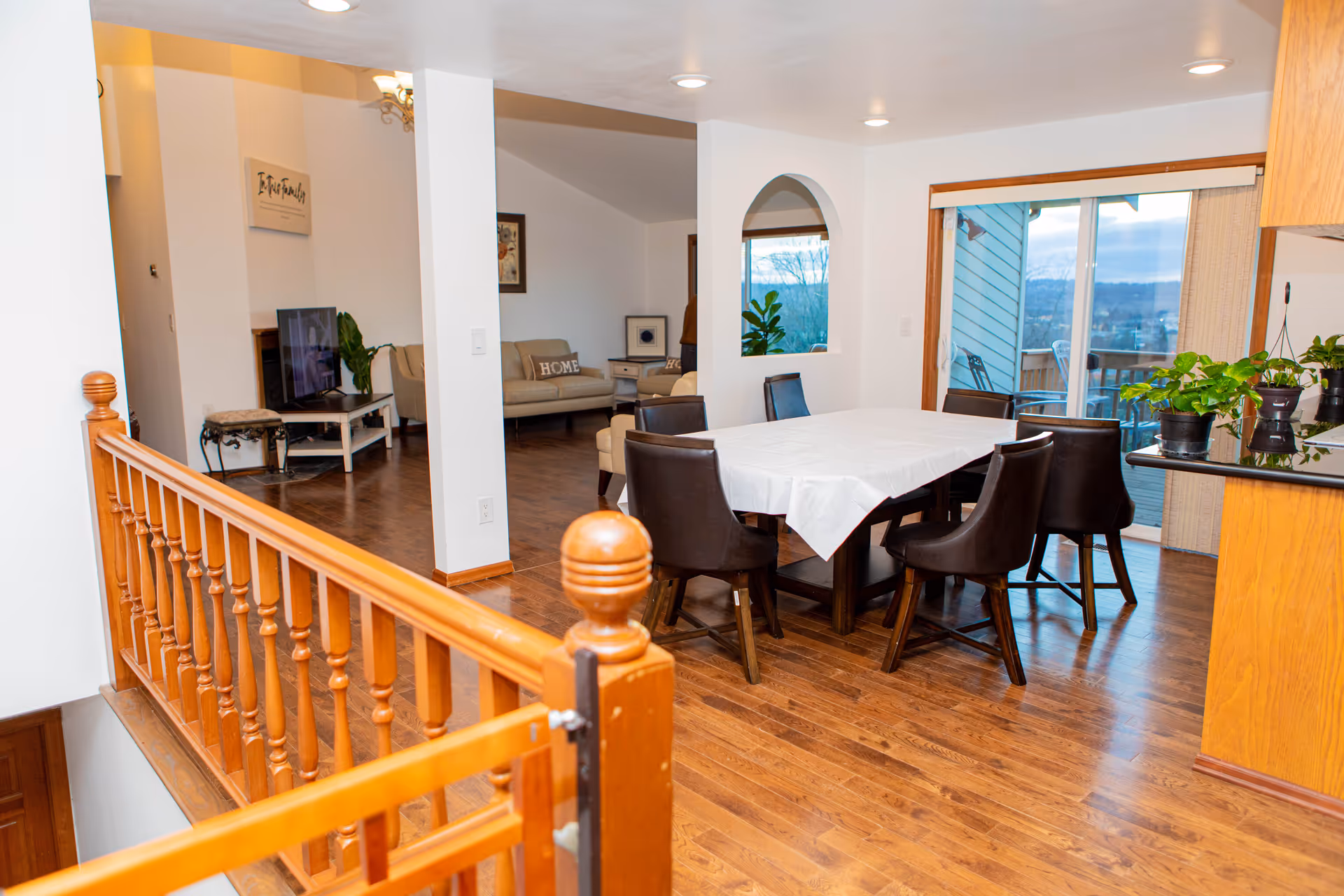 Interior view of a senior living facility showing a dining area with a table covered by a white tablecloth and six dark chairs. Adjacent to the dining area is a living room with beige sofas, a TV on a stand, and decorative wall art. The space has wooden flooring, a wooden railing, and a sliding glass door leading to an outdoor balcony with a view.