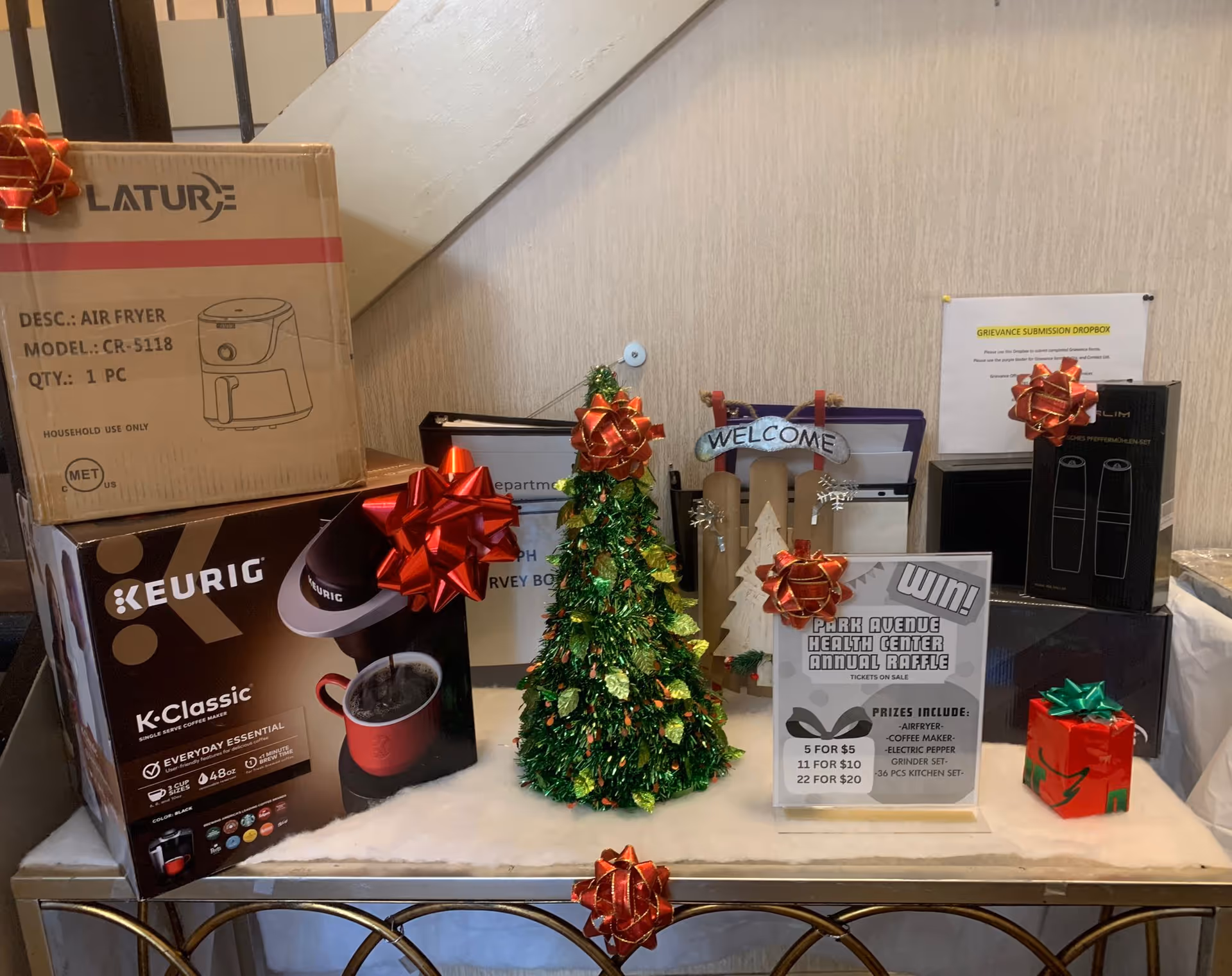 Holiday raffle display on a table inside a facility featuring a small decorated Christmas tree, wrapped gifts and boxed prizes including a Keurig and an air fryer, with a 'Welcome' sign and a Park Avenue Health Center raffle poster.