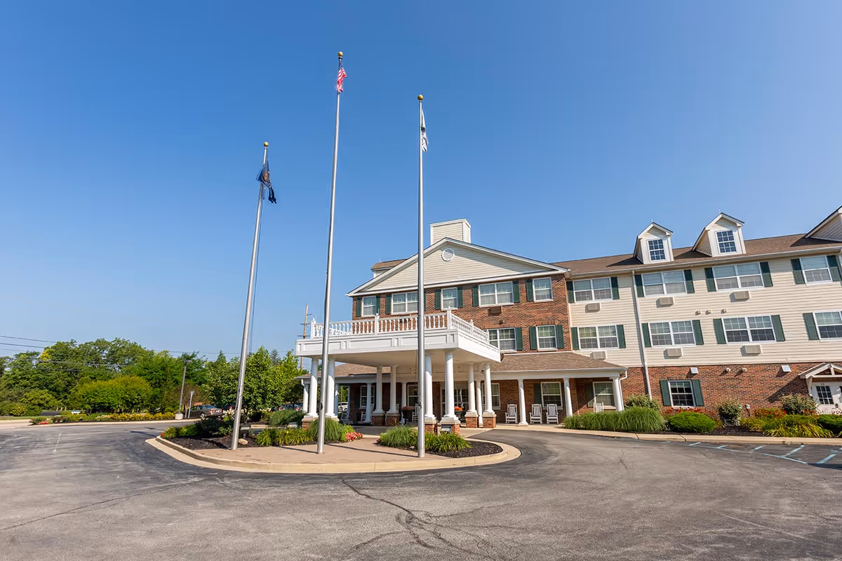 Exterior view of the Grand Victorian of Zionsville senior living facility showing a large three-story building with a covered entrance supported by white columns, three flagpoles with flags, and surrounding landscaping under a clear blue sky.