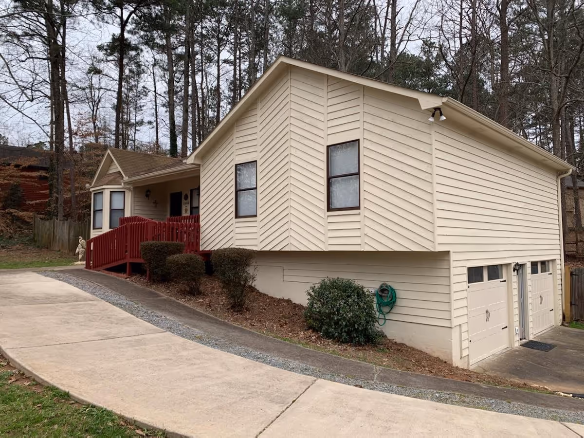Exterior view of a beige single-story house with a sloped driveway, red wooden ramp leading to the front door, two garage doors, and surrounded by trees and bushes.