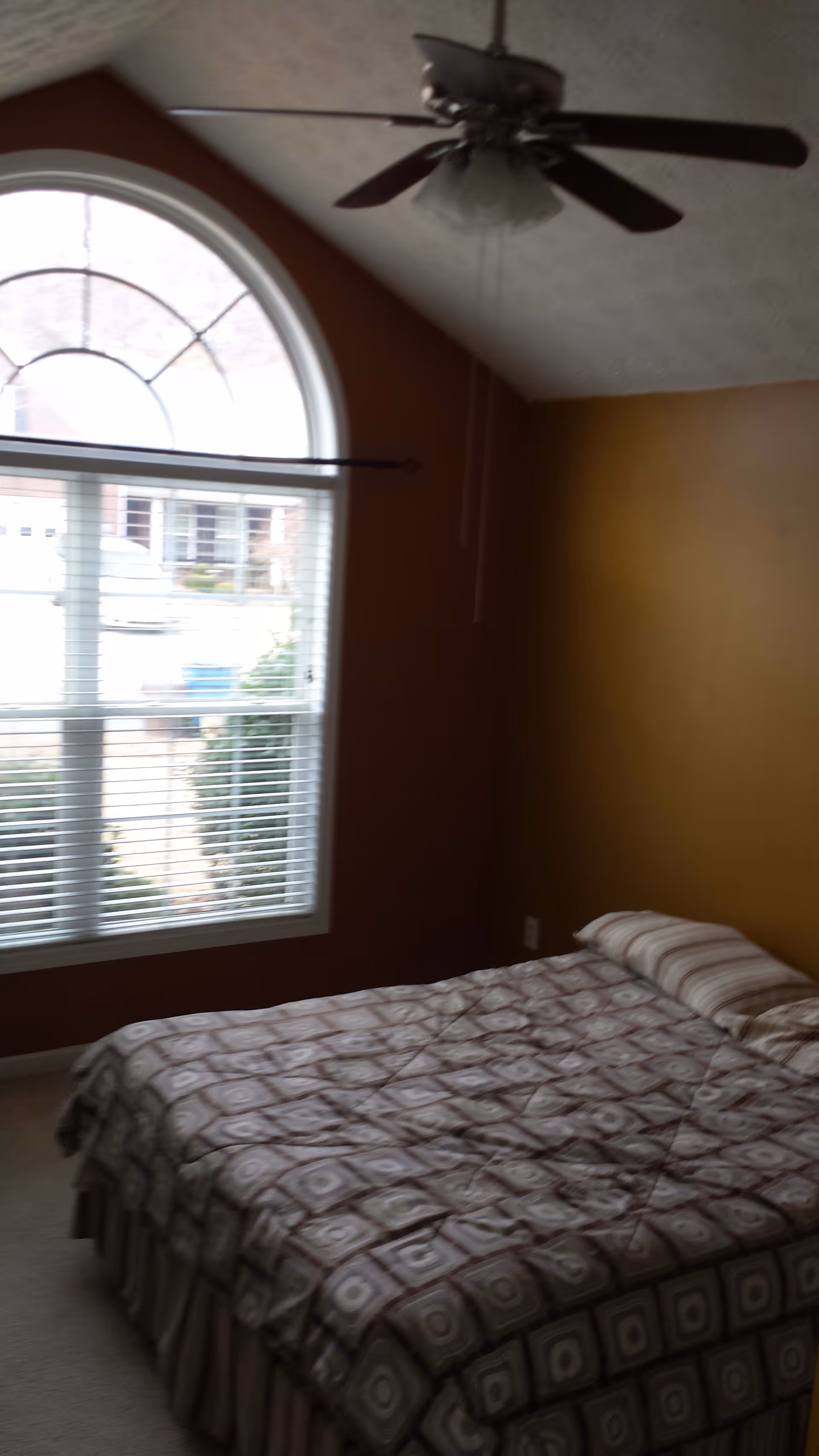 A simple bedroom with a bed covered in a patterned comforter, an arched window with blinds, and a ceiling fan.