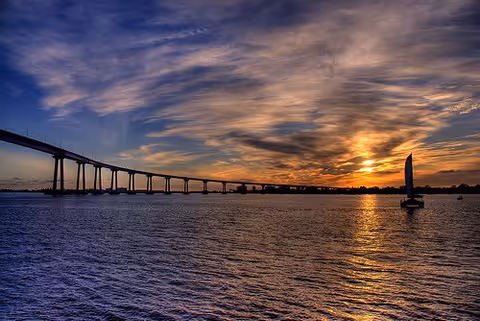 A long bridge extending across a body of water during sunset with a sailboat on the right side and a dramatic sky filled with clouds.